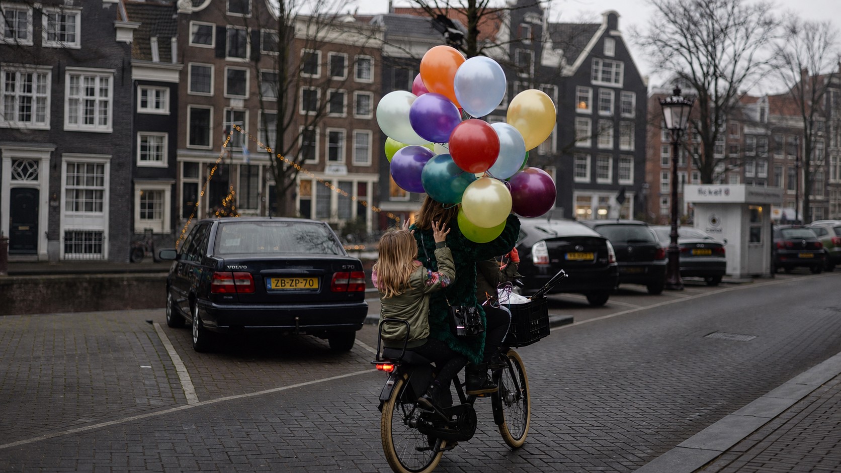 A woman cycling along an Amsterdam canal with a cluster of balloons, a child riding behind her